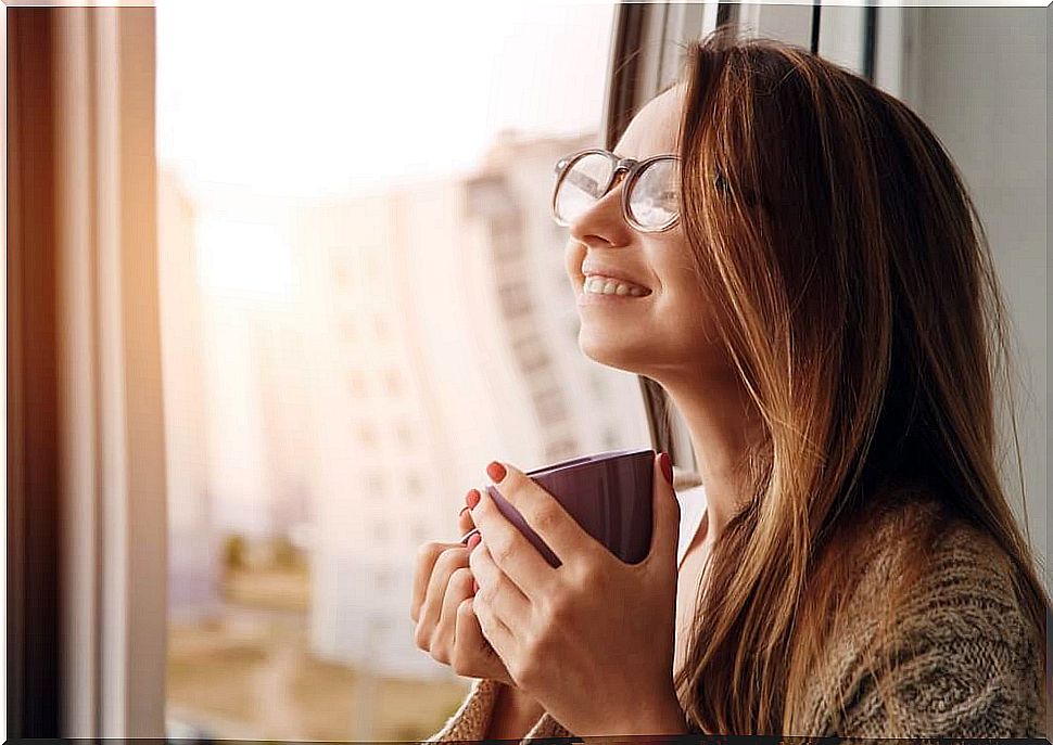 Woman with a mug in her hand smiling and looking out the window.